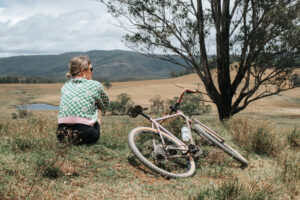 Rider resting on a grassy hilltop beside her bike on the Timbarra–Lindrook Loop